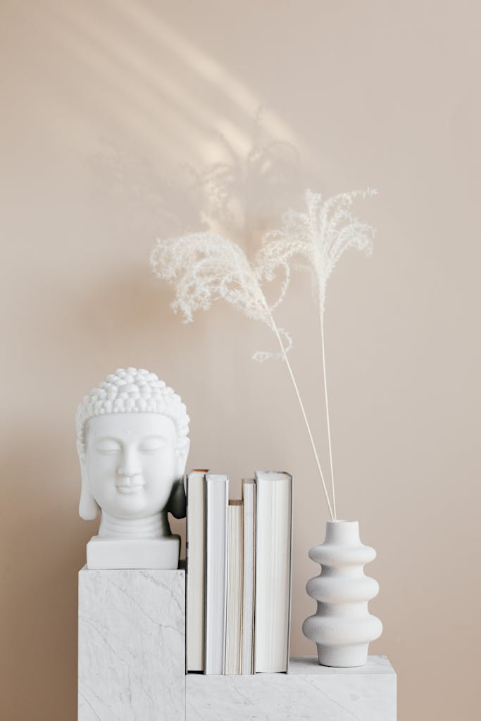 A minimalist decor setup featuring a Buddha head sculpture with books and a vase on a marble shelf.